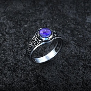 May include: A silver-toned ring featuring a round, faceted purple gemstone set in the center. The band is decorated with an intricate, repeating knotwork pattern that extends around the sides of the ring, contrasting with the dark metal finish.