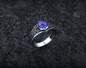 May include: A silver-toned ring featuring a round, faceted purple gemstone set in the center. The band is decorated with an intricate, repeating knotwork pattern that extends around the sides of the ring, contrasting with the dark metal finish.
