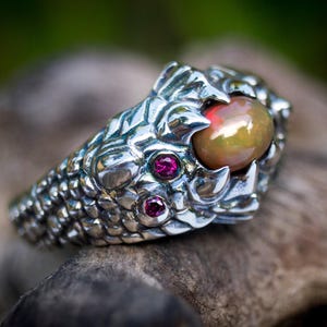May include: A silver ring featuring a detailed dragon head design with textured scales. The dragon's eyes are set with two small, round, vibrant red gemstones, and it holds a smooth, oval-shaped iridescent opal cabochon in its open mouth.