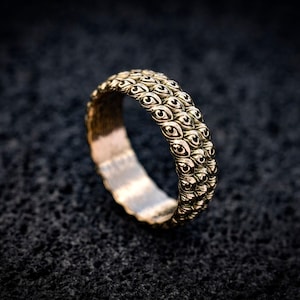 May include: A metallic ring featuring a repeating pattern of small, sculpted eyes covering the entire outer surface. The band has a textured, dark finish with polished highlights on the raised eye details, creating a distinct, repetitive visual motif.