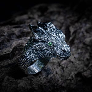 Op de afbeelding: Een zilverkleurige metalen ring in de vorm van een drakenkop met gedetailleerde schubstructuren en een enkele kleine, felgroene edelsteen in de oogkas. De ringband is voorzien van een bijpassend schubpatroon over het gehele oppervlak.