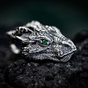 May include: A silver dragon head ring with a green gemstone eye. The ring is detailed with scales and a textured surface.
