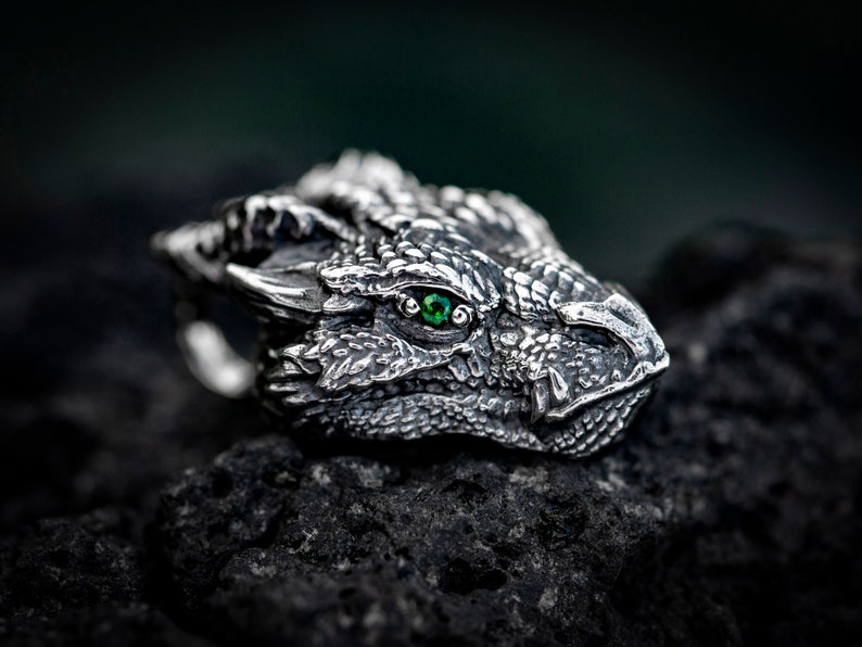 May include: A silver dragon head ring with a green gemstone eye. The ring is detailed with scales and a textured surface.