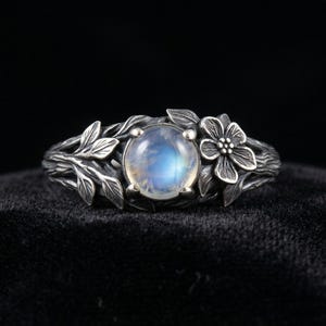 Könnte beinhalten: Ein Silberring mit einem zentralen, runden Mondstein, der einen blauen, irisierenden Schimmer aufweist. Die Ringschiene ist mit detaillierten, oxidierten Silberstrukturen gestaltet, die an Äste erinnern, verziert mit Blättern und einer Blume.