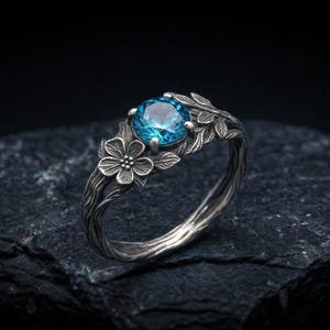 May include: A silver ring featuring a round, faceted blue gemstone set in the centre. The band is designed to resemble twisted tree branches and is adorned with detailed metal leaves and a small five-petaled flower on one side of the stone.
