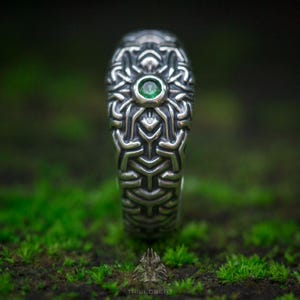 May include: A silver ring featuring an intricate, raised geometric pattern across its surface. A single, small, round, vibrant green gemstone is set centrally within the metalwork. The ring has a polished finish with darkened recessed areas.