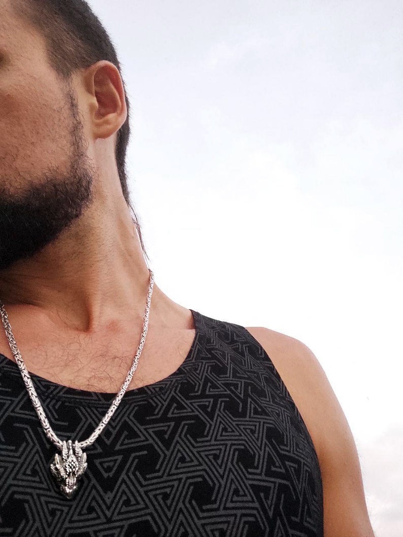 May include: A silver chain necklace with a dragon head pendant. The necklace is worn by a person wearing a black tank top with a geometric pattern.
