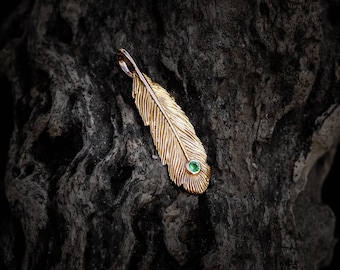 May include: A gold feather pendant featuring a detailed, textured surface that mimics the appearance of natural plumage. A small, round, vibrant green gemstone is set near the bottom of the feather, adding a distinct point of color to the piece.