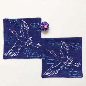 Puede incluir: Dos posavasos de lino azul con grullas bordadas en blanco volando sobre un fondo de nubes azules. Los posavasos son cuadrados y tienen un borde decorativo. Un pequeño adorno decorativo redondo, morado y rosa, se encuentra en la esquina superior derecha de la imagen.