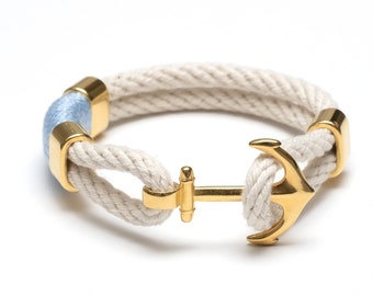May include: A white rope bracelet with a gold anchor charm and a light blue accent. The bracelet is made of two strands of rope and has a gold clasp.