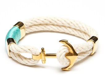 May include: A white rope bracelet with a gold anchor charm and a turquoise blue accent. The bracelet is made of two strands of rope and has a gold clasp.