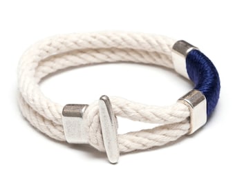 May include: A nautical-themed bracelet featuring a thick, off-white rope design. The bracelet has a dark blue accent and silver-colored metal clasps. The rope is approximately 0.5 inches thick, and the bracelet is designed to be worn around the wrist.