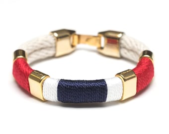May include: A red, white, and blue rope bracelet with gold accents. The bracelet is made of braided rope and has a gold clasp.