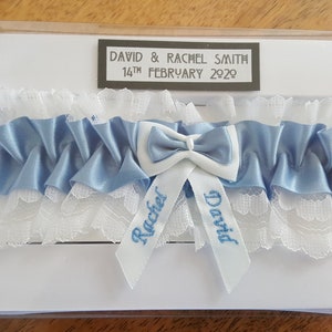May include: A white and blue garter with the names "David" and "Rachel" embroidered on it. The garter is in a clear plastic box with a white card that says "David & Rachel Smith, 14th February 2020".