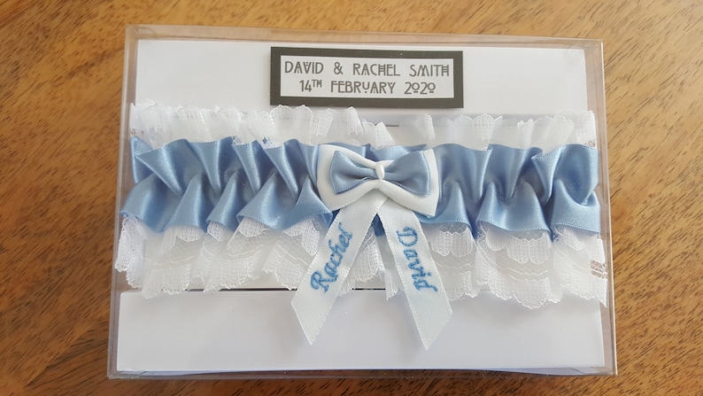 May include: A white and blue garter with the names "David" and "Rachel" embroidered on it. The garter is in a clear plastic box with a white card that says "David & Rachel Smith, 14th February 2020".