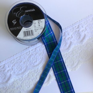 May include: A roll of blue and green plaid ribbon with a spool of black ribbon in the background. The plaid ribbon is 10 inches wide and 25 yards long. The spool of black ribbon is labeled "essentials" and has the text "Berisfords" on it.