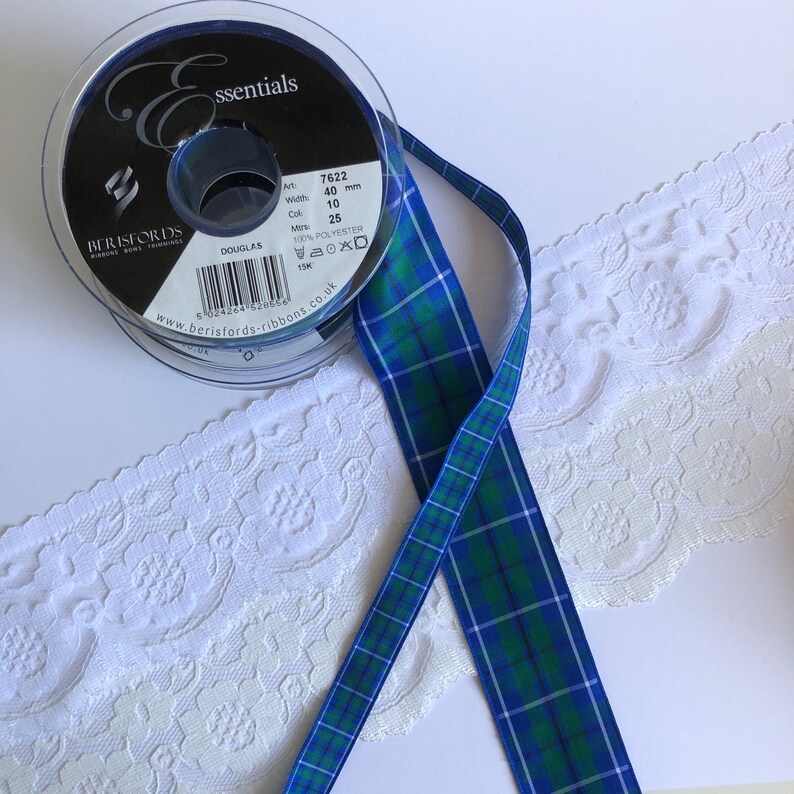 May include: A roll of blue and green plaid ribbon with a spool of black ribbon in the background. The plaid ribbon is 10 inches wide and 25 yards long. The spool of black ribbon is labeled "essentials" and has the text "Berisfords" on it.