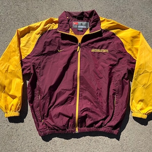 Op de afbeelding: Bordeauxrode en gouden Nike Arizona State jas. De jas heeft een volledige ritssluiting, een opstaande kraag en de woorden "ARIZONA STATE" zijn op de borst geborduurd. De mouwen zijn goudkleurig en de body is bordeauxrood. Het Nike-logo is zichtbaar.