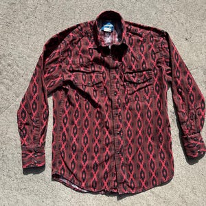 Op de afbeelding: Een shirt met lange mouwen in een rood en zwart geometrisch patroon. Het shirt heeft twee borstzakken en manchetten met knopen. De kraag heeft een blauw-wit label.