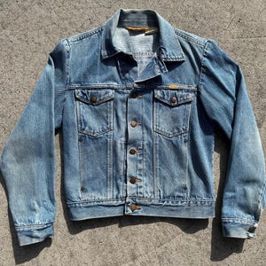 May include: A light blue denim jacket with a button-up front, two chest pockets, and two side pockets. The jacket has a classic collar and long sleeves. The denim has a faded appearance.