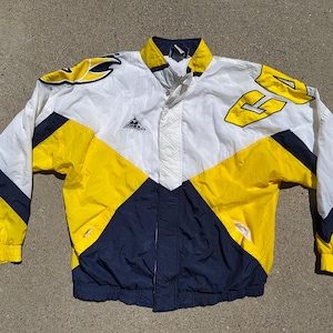 Könnte beinhalten: Eine Vintage Apex Jacke in Weiß, Gelb und Marineblau. Die Jacke hat ein Colorblock-Design mit gelben Akzenten an den Ärmeln und am Körper. Die Jacke hat eine Knopfleiste vorne und einen Kragen. Auf der Brust befindet sich ein Logo.