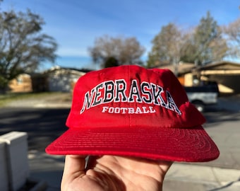 May include: Red baseball cap with white embroidered text that reads "Nebraska Football".