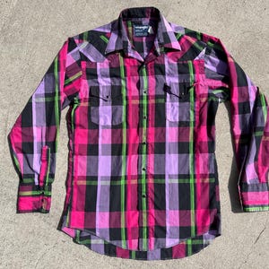 Op de afbeelding: Een button-down shirt met lange mouwen met een ruitpatroon in roze, zwart, groen en paars. De shirt heeft twee borstzakken en een kraag. Het Wrangler-merklabel is zichtbaar op de kraag.