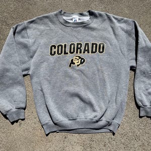 Może przedstawiać: Szara bluza z okrągłym dekoltem z napisem "COLORADO" w czarnych i złotych literach na piersi. Poniżej czarno-złote logo. Bluza ma długie rękawy oraz ściągacze przy mankietach i kołnierzu.