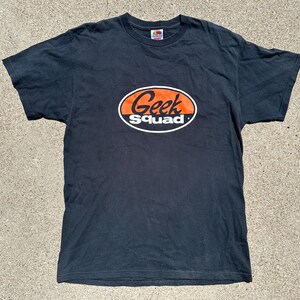 May include: Black short-sleeved t-shirt with the words "Geek Squad" in an orange and white oval design on the front. The shirt is made of a soft material and has a crew neck.