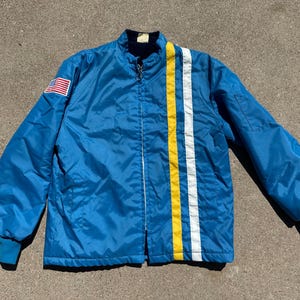 Op de afbeelding: Een vintage blauwe jas met ritssluiting, met verticale gele en witte strepen. Een Amerikaanse vlag patch staat op de linkermouw. De jas heeft een opstaande kraag en elastische manchetten.