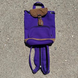 May include: A purple backpack with a brown suede flap and pocket. The backpack has adjustable straps and a top handle. The bag has a front pocket and a drawstring closure. The backpack is made of a durable material.