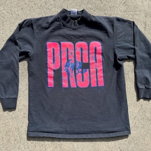 Op de afbeelding: Donkergrijs shirt met lange mouwen en een opstaande kraag. Het shirt heeft het woord "PRCA" in grote, roze blokletters over de borst, met een blauwe afbeelding van een rodeo-scène in de letters. Het shirt is vintage.