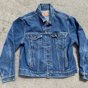 May include: A classic blue denim jacket with a button-up front, two chest pockets, and two side pockets. The jacket has a collared neckline and buttoned cuffs. The denim is a medium wash, and the jacket appears to be vintage.