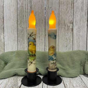 Könnte beinhalten: Zwei gelbe LED-Kerzen mit einem Vogel- und Blumendesign an der Seite. Die Kerzen stehen auf schwarzen Kerzenständern.