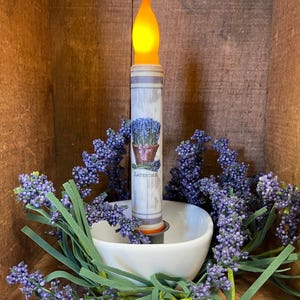 May include: A white pillar candle with a lavender design and the word "Lavender" printed on it. The candle is lit and has a yellow flame. The candle is surrounded by purple lavender sprigs.