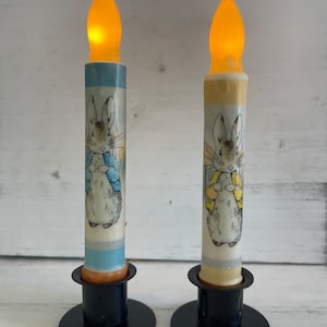 Puede incluir: Dos portavelas a pilas con un diseño de Peter Rabbit. Las velas son blancas con rayas azules y amarillas y presentan una ilustración de Peter Rabbit. Los portavelas son de metal negro.