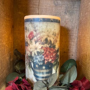 Könnte beinhalten: Zylindrische Kerze mit floralem Design in Rot, Weiß und Blau, in einer patriotischen Vase. Künstliches Grün und rote Blumen umgeben die Kerze, vor einem Holz Hintergrund platziert.