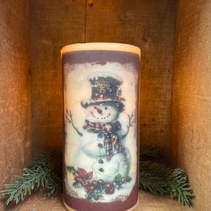 May include: A cylindrical, battery-operated candle featuring a snowman design. The snowman wears a black top hat, a plaid scarf, and has twig arms. The candle has a burgundy border and is set against a rustic wooden background. Decorative greenery is placed around the candle.