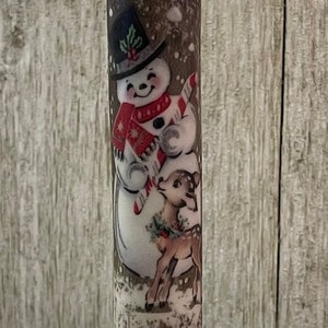 Puede incluir: Una vela navideña a pilas con un diseño de muñeco de nieve y ciervo. La vela tiene una base roja y un portavelas negro. La llama es de color amarillo cálido. Una decoración festiva.