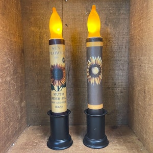 May include: Two black candle holders with a yellow flame on top. Each candle has a brown and yellow sunflower design with the text "SUN FLOWER REG. HUTH SEED CO. SAN ANTONIO TEXAS".