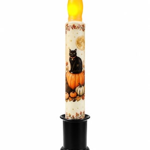 Può includere: Una candela affusolata a batteria con una punta gialla a forma di fiamma, caratterizzata da un corpo color crema decorato con un gatto nero seduto su una zucca arancione, circondato da zucche più piccole, foglie autunnali e una luna piena, su base nera.