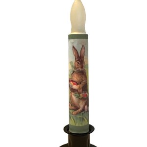May include: A white candle with a brown rabbit and eggs design on the side. The candle is on a black metal candle holder.