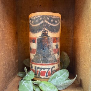 Puede incluir: Una vela pilar con un diseño de Campana de la Libertad rojo, blanco y azul y el año 1776 impreso en el lateral. La vela está en una caja de madera con follaje verde.