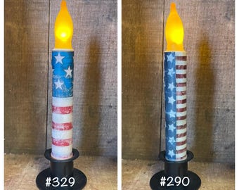 May include: Two decorative candles with a red, white and blue American flag design. The candles are on black candle holders with the numbers #329 and #290 printed on them.