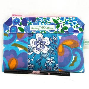 May include: A rectangular, zippered pouch with a patchwork design. The pouch features a pink zip and is adorned with floral patterns in shades of blue, purple, and orange. The text "You're a Saucy Old Minx" is visible on a label.