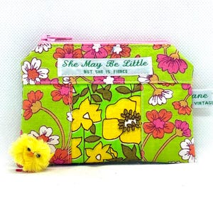 May include: A small, zipped coin purse with a bright floral pattern in green, pink, orange, and yellow. The purse has the text "She May Be Little But She Is Fierce." A small, yellow chick is next to the purse.