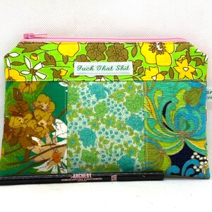 May include: A patchwork zipped pouch with a pink zip and various floral patterns in green, yellow, and blue. The pouch has the text "Fuck That Shit" on a label. A black eyeliner pencil is in front of the pouch.