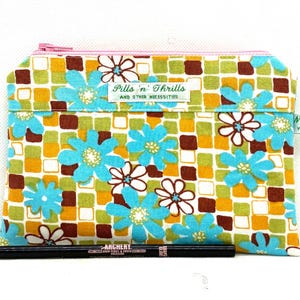 May include: A rectangular fabric zip pouch featuring a pattern of turquoise and white flowers over a grid of green, yellow, and brown squares. A white label reads "Pills 'n' Thrills AND OTHER NECESSITIES". A black eyeliner pencil lies in front.