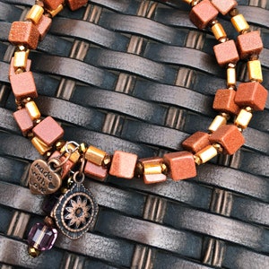 May include: A double-strand bracelet made with brown and gold square beads. The bracelet has a charm with the words "Made with Love" and a floral design.