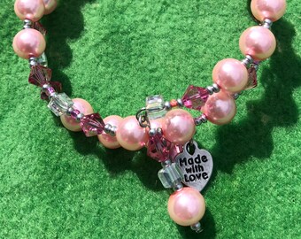 Könnte beinhalten: Ein rosa Perlenarmband mit einem silbernen Anhänger, auf dem "Made with Love" steht. Das Armband ist aus kleinen rosa und klaren Kristallen gefertigt.